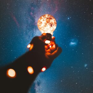 A hand holds a glowing bulb into the Milky Way night sky, creating a magical cosmic connection.