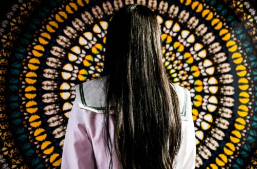 žena s dlouhými tmavými vlasy stojí před živým pozadím s mandalovým vzorem, což vytváří výrazný vizuální kontrast.