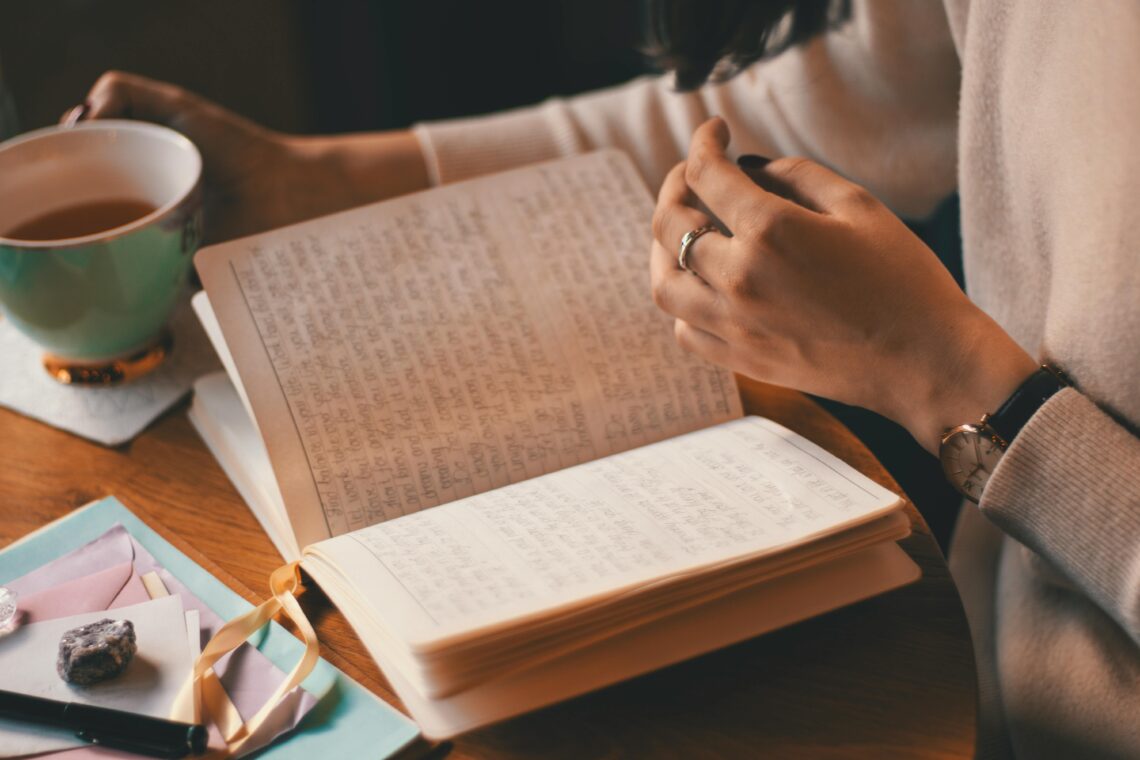 Žena píše do deníku, zatímco si vychutnává šálek kávy u dřevěného stolu.