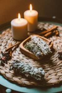 A warm and inviting scene with candles, sage bundles, and cinnamon sticks on a woven mat.