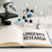 Fotografie laboratorního stolu s mikroskopem, molekulovým modelem, laboratorním sklem a otevřenou knihou s nápisem Longevity Research – výzkum dlouhověkosti“- Photo of a laboratory table with a microscope, molecular model, laboratory glassware, and an open book with the title "Longevity Research."