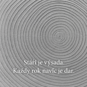 Detailní černobílá fotografie letokruhů dřeva. Viditelné soustředné kruhy symbolizují roky a růst. V obrázku je umístěný nápis „Stáří je výsada. Každý rok navíc je dar.“