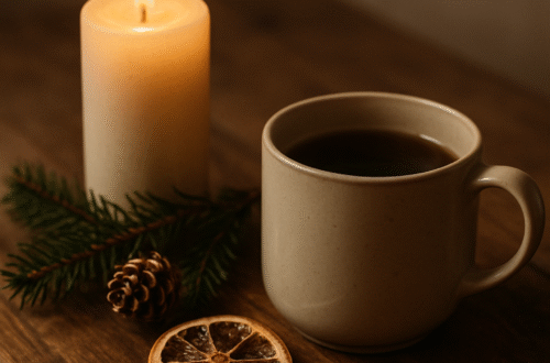 Teplý hrnek čaje a zapálená svíčka na dřevěném stole, doplněné o šišku a větvičku jehličí. Přírodní zimní atmosféra v klidných, hřejivých barvách.
