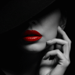 Detailní černobílý portrét ženy s kloboukem, který zakrývá horní část obličeje. Dominantou jsou výrazné, lesklé červené rty. Minimalistický, elegantní, ženský styl.