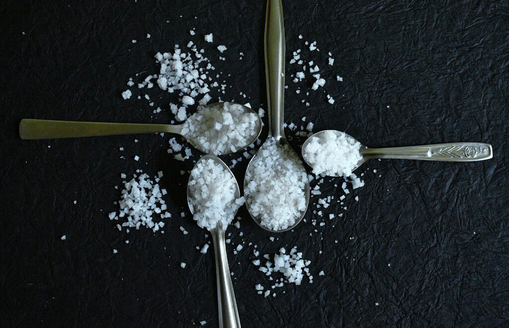 Overhead view of coarse sea salt on silver spoons against a textured black background.