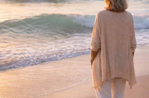 Žena po šedesátce stojí bosá na pláži při východu slunce a dívá se na dvě přicházející vlny – symbol přirozených fází stárnutí.