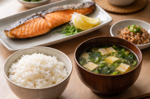 Minimalistický japonský stůl s miskou bílé rýže, miso polévkou s tofu, grilovaným lososem a šálkem zeleného čaje v přirozeném denním světle.