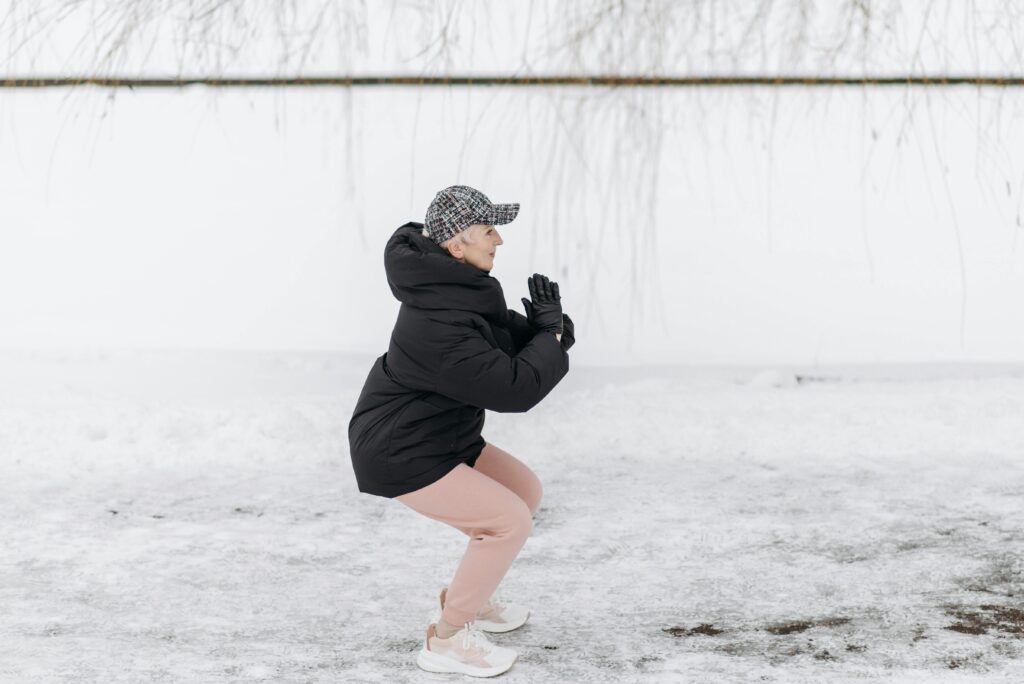 zdravy pohyb, svaly, Senior woman doing squats outside in winter clothing on snowy ground.