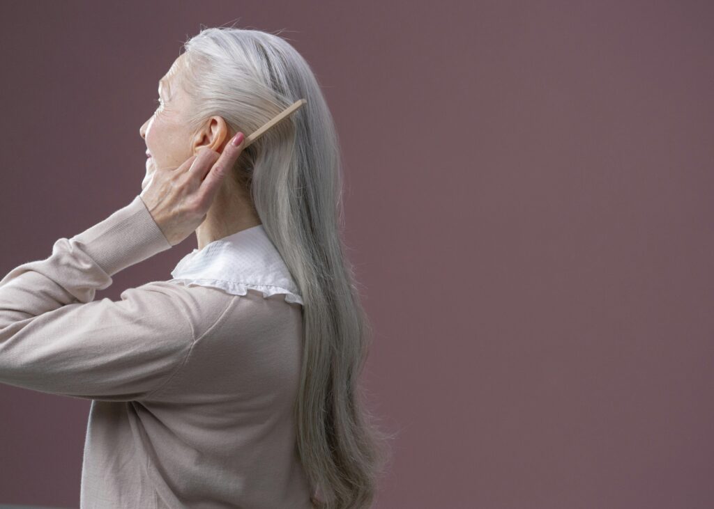 šedivé vlasy, krása, stříbro ve vlasech, Elderly woman with long grey hair combing it, set against a purple background.
