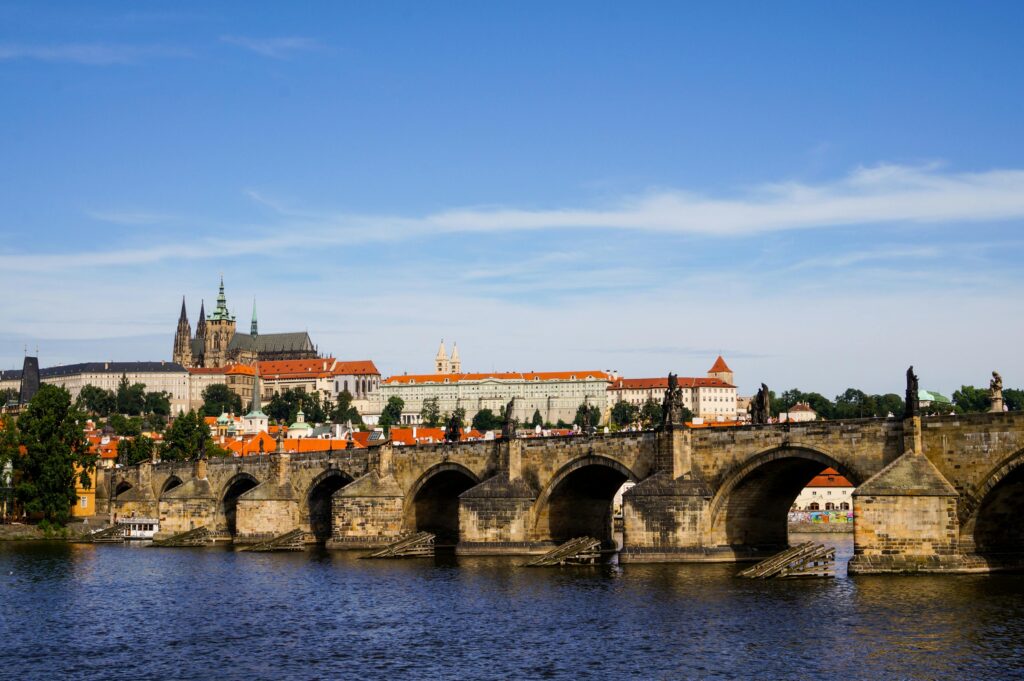 A možná je čas se zeptat: patřit kam, vlastně? A za jakou cenu? View of Charles Bridge with Prague Castle in the background on a sunny day.