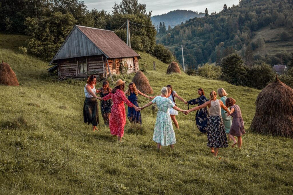 keltské kolo roku, Lammas, oslavy dožínky, Celtic Wheel of the Year, A group of women dancing in a rural meadow beside a wooden hut, surrounded by nature.